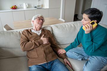 Concerned wife makes urgent call during husband heart attack. Senior male collapsing, chest pain, panic in room, aging health risk, cardiovascular failure, immediate assistance and domestic emergency