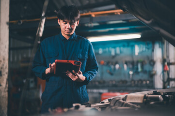 A mechanic in a blue jumpsuit uses a tablet to diagnose or inspect a vehicle in a workshop with...