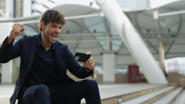 Portrait of emotional Hispanic businessman watches favorite sports game via phone and sits on steps. Latin man expresses excitement of goal in match raising fist shouting in city