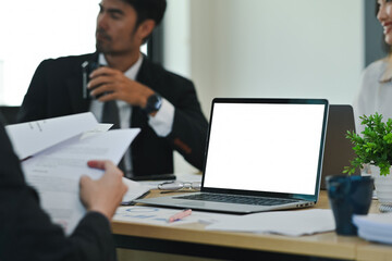 Close-up of a workspace with a blank laptop screen and paperwork during a corporate team meeting