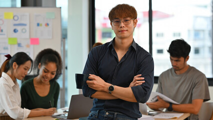 Handsome Team leader poses proudly as colleagues engage in discussion and data analysis in a modern office