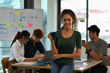 Confident young African woman holding a coffee mug while standing in a modern office, team members working in the background