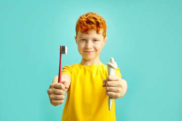 Red haired little boy holding brush tooth and tube of dental paste, preparing to clean teeth,...