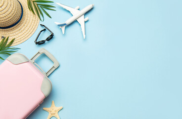 Pink suitcase, airplane model, sunglasses and straw hat on blue background with copy space
