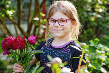 Cute little school girl with eyeglasses holding blooming dark pink peonies in garden. Happy child...