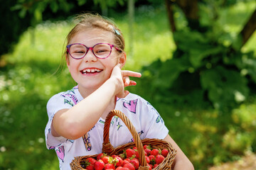 Happy little school girl holding a big basket full of ripe, sweet strawberries. Real life, real people moment of childhood joy and summer fun outdoors in nature.