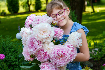Close-up of little school girl holding a large bunch of blooming pink peonies in a garden. Happy child enjoying a spring or summer day outdoors. Real life, real people, natural childhood moment