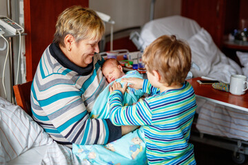 Happy grandmother holding newborn baby in hospital with proud big brother nearby. Real life, real people moment of family love, siblings, and multigenerational bonding.