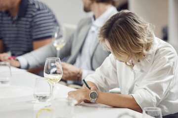 Female member of sommelier school tasting white wine and makes notes in degustation card. Wine expert studies alcoholic drink. 