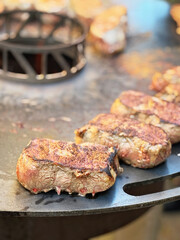 Many pieces of juicy beef steaks sizzling on a barbecue grill. Fresh meat cooking outdoors for a summer BBQ. Real life, real people, food preparation and grilling concept