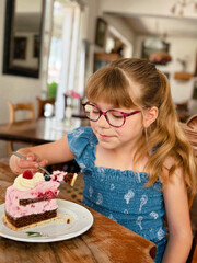 Little girl with eyeglasses eating a big piece of berry cake in a cozy cafe. Real life, real people moment of childhood joy, sweet treat, and delicious dessert enjoyment.