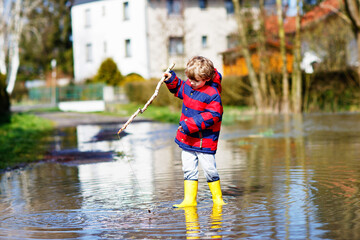 Little kid boy wearing yellow rain boots and walking and jumping into puddle on warm sunny spring day.