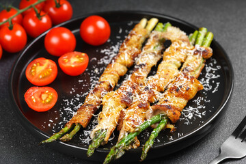 Grilled asparagus wrapped in crispy bacon sprinkled with parmesan cheese served on plate with fresh cherry tomatoes on black background. Gourmet dish, cooking recipe, cuisine, or restaurant menu