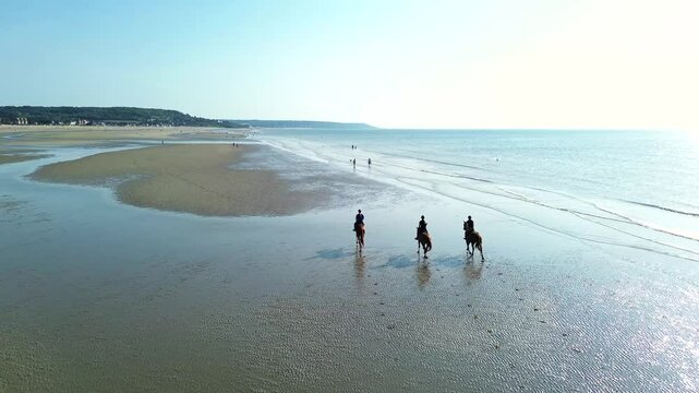 Drone shots of riders galloping at full speed across the reflective shoreline of Deauville beach. The sun glistens on the water as hooves kick up spray,