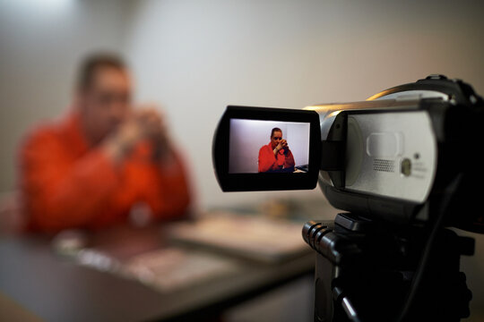 Caucasian middle aged man sitting at table in prison uniform being filmed by video camera, hands clasped near face, focus on camera display showing subject in frame