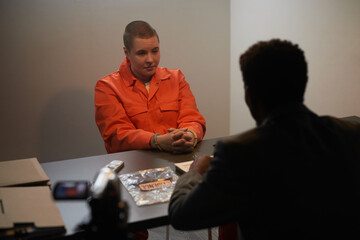Caucasian young adult woman in orange prison jumpsuit sitting handcuffed at table, facing Black...