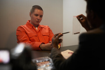 Caucasian young adult woman with short hair wearing orange prison jumpsuit, sitting handcuffed at...