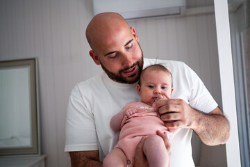 A man is holding his infant child in a bright, cozy room. The baby is dressed in light pink...