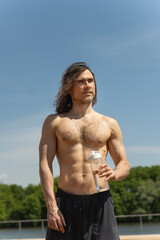 Shirtless fit brunette man with curly long hair holding water bottle looking away against clear blue sky and nature background. Heat summer.