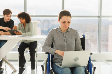 Accessible environment. Woman in chair for people with disability working with laptop in modern office. Diverse multiracial group of people colleagues working together. Young professional team
