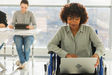 Accessible environment. African woman in chair for people with disability working with laptop in modern office. Diverse multiracial group of people colleagues working together. Young professional team