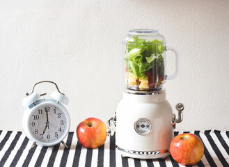 white vintage blender or smoothie maker machine  with vegetables and fruit inside, white vintage alarm clock shows 7 o'clock and 2 apples  on  black and white stripe table cloth and white wall.