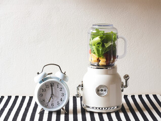 white vintage blender or smoothie maker machine  with vegetables and fruit inside and white vintage alarm clock shows 7 o'clock  on  black and white stripe table cloth and white wall.