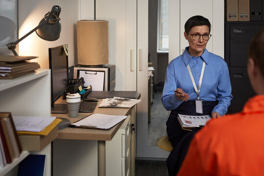 Middle aged Caucasian woman psychologist conducting counseling session with prisoned young adult in orange uniform, sitting in office, holding pen and discussing documents
