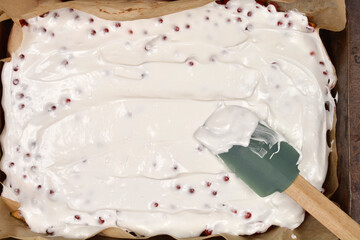 A silicone spatula finishes spreading a smooth, white meringue topping over a red currant cake in a...