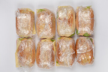 Eight individual sandwiches, each neatly wrapped in clear plastic film, are arranged in two rows on a clean white surface, prepared for a convenient meal