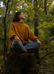 Young Caucasian woman enjoying autumn nature and weather sitting on a tree stump in autumn forest