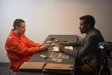 Caucasian teenager with shaved head wearing orange prison uniform, sitting at table facing Black...