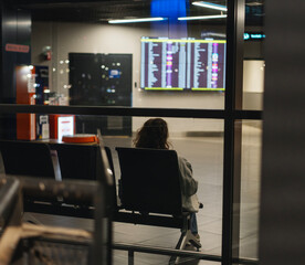 Woman sitting on bench in front of flight schedule at night airport