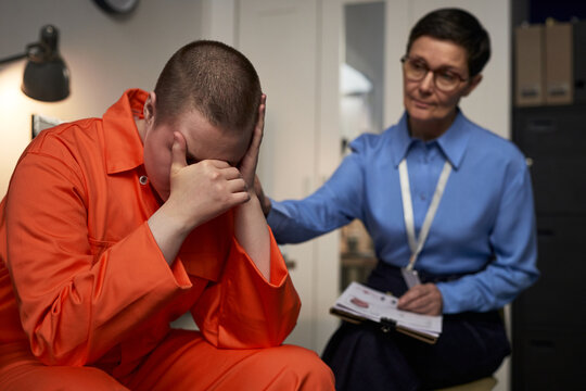 Caucasian young adult woman in orange prison uniform sitting with head in hands, showing distress while middle aged Caucasian woman counselor offering support during prison visit