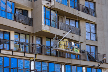 Construction worker on suspended platform performs facade plastering on tall urban building,...