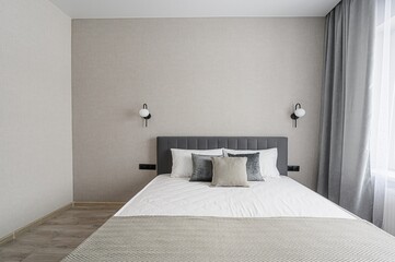 serene bedroom with a gray headboard bed, white linens, and neutral-toned walls. Symmetrical wall lamps frame the bed. Curtains filter light