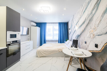Modern studio apartment with marble-effect wallpaper, blue curtains, a bed, and a kitchen area. White marble floor