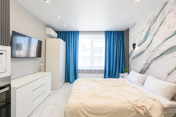well-lit modern bedroom with a bed, blue curtains, a TV, and white furniture. A decorative marble-patterned wall adds a stylish touch