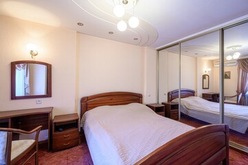 Cozy bedroom featuring a wooden bed, mirrored closet, desk, and warm lighting. Peach-colored walls and carpet create a warm atmosphere