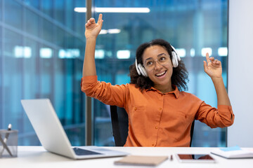 A joyful young woman wearing headphones and glasses, celebrating in an office setting with a laptop.