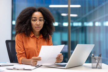 A serious woman with curly hair wears glasses and examines paperwork, with a laptop on a desk in an...