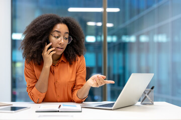 A woman with glasses, speaking on the phone while working on a laptop at her desk. She appears...