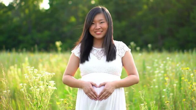 a young woman making a heart shape with her hands on top of her belly outdoors