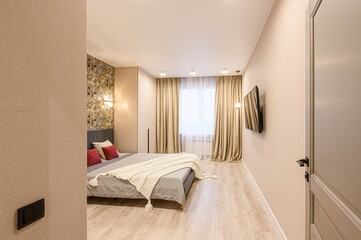 cozy bedroom features a bed with red pillows, light wood floors, beige walls, and a TV. Natural...