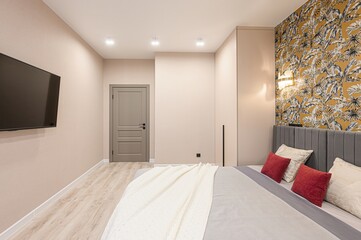 modern bedroom with beige walls, wood floor, and a bed with red pillows. A TV is mounted on the wall opposite a door. Wallpaper with a palm leaf print is on the back wall