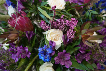 Vibrant floral bouquet with white roses, purple alstroemeria, pink protea, blue delphinium, pink statice, succulent-type foliage, and green leaves.