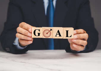 Businessman holding wooden blocks spelling goal with dartboard icon symbolizing achievement.