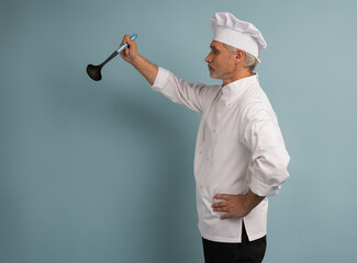 Mature male chef with spoon tasting dish on light blue background.