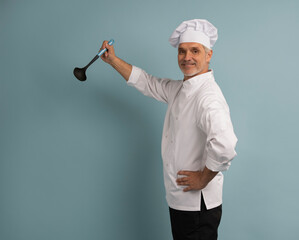Mature male chef with spoon tasting dish on light blue background.