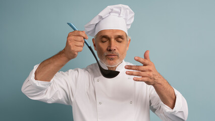 Mature male chef with spoon tasting dish on light blue background.